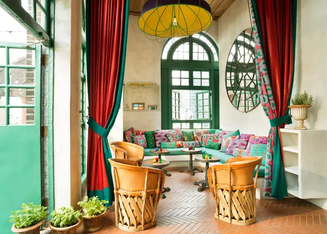 A lounge corner with a teal banquette covered in patterned cushions, wicker armchairs, round tables, red and green drapes and a yellow pendant lamp.