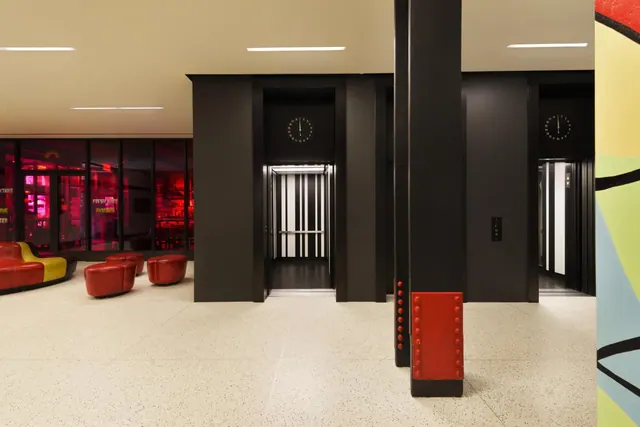 A lobby with two lift entrances in black walls, a black pillar with a red base, red ottoman seats and a red and yellow sofa beside a glass-fronted area.