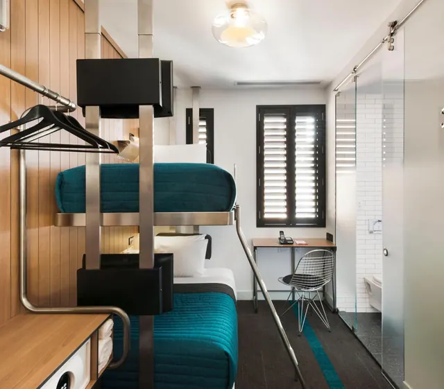 A hotel room with teal-covered bunk beds, a wooden wall, a small desk with a telephone and a wire chair, and a glass-walled bathroom on the right.