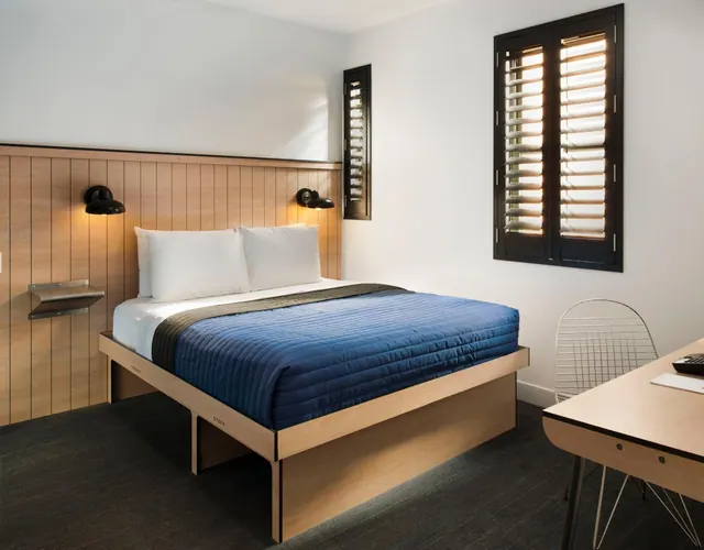 A bedroom with a double bed under a blue quilt, a wood-panelled headboard wall, wall lamps, and a desk beside a shuttered window.