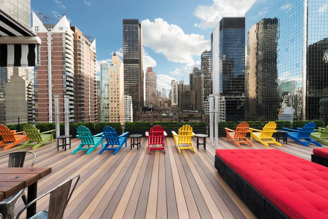 A rooftop terrace with wooden decking holds rows of brightly coloured Adirondack chairs and a red cushioned daybed, with tall city towers behind.