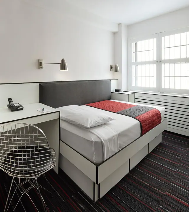 A bedroom with a double bed under a red and grey throw, a built-in desk with a telephone and a wire chair, and a large window with white shutters.