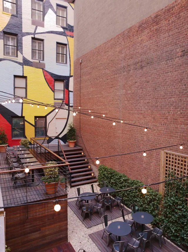 An outdoor courtyard with wooden decks, metal tables and chairs, strung lights, a hedge, and a colourful painted mural on the building wall.
