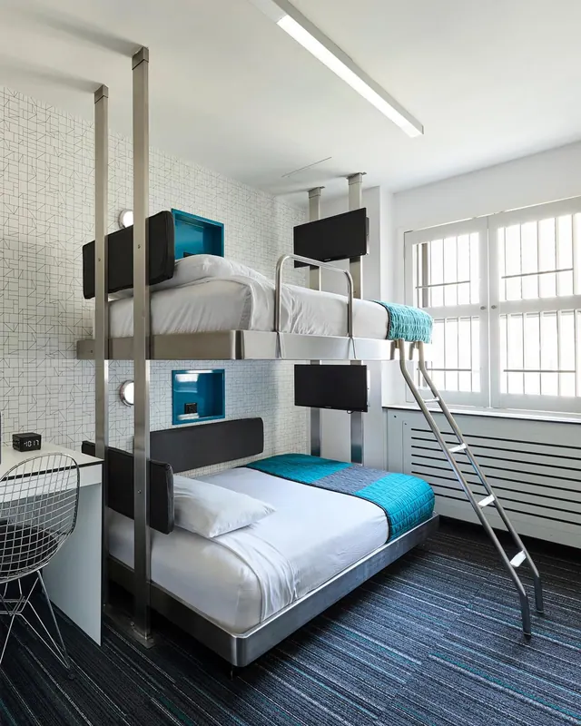A bedroom with a metal bunk bed frame, a double bed below with a ladder, a wire chair at a desk, and a window with shutters.