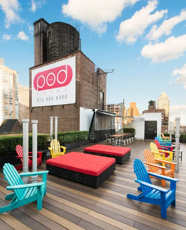 A rooftop terrace with brightly colored Adirondack chairs and red cushioned ottomans, with a Pod Hotel sign on a brick building and city buildings behind.
