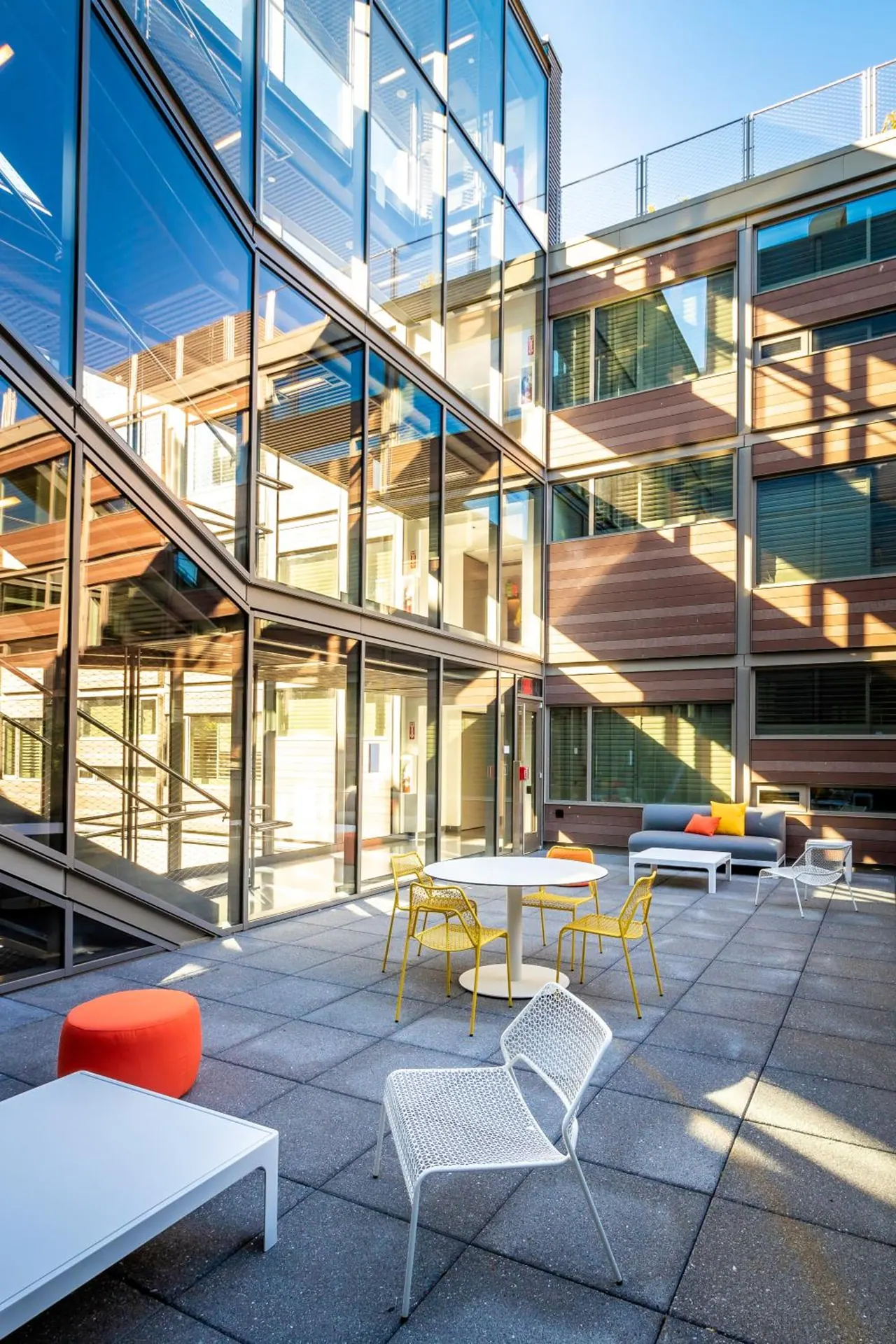 A paved outdoor courtyard with white and yellow chairs, a round table, a grey sofa and a glass-fronted building around it.