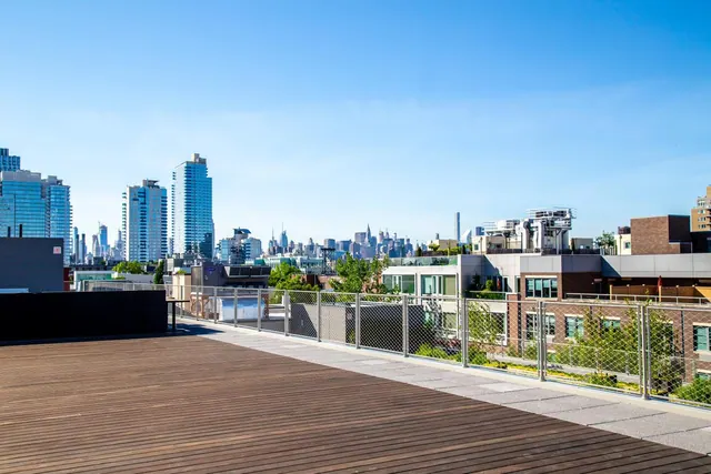 A wooden-decked rooftop terrace with a metal mesh railing overlooks neighbouring buildings and a city skyline under a clear blue sky.