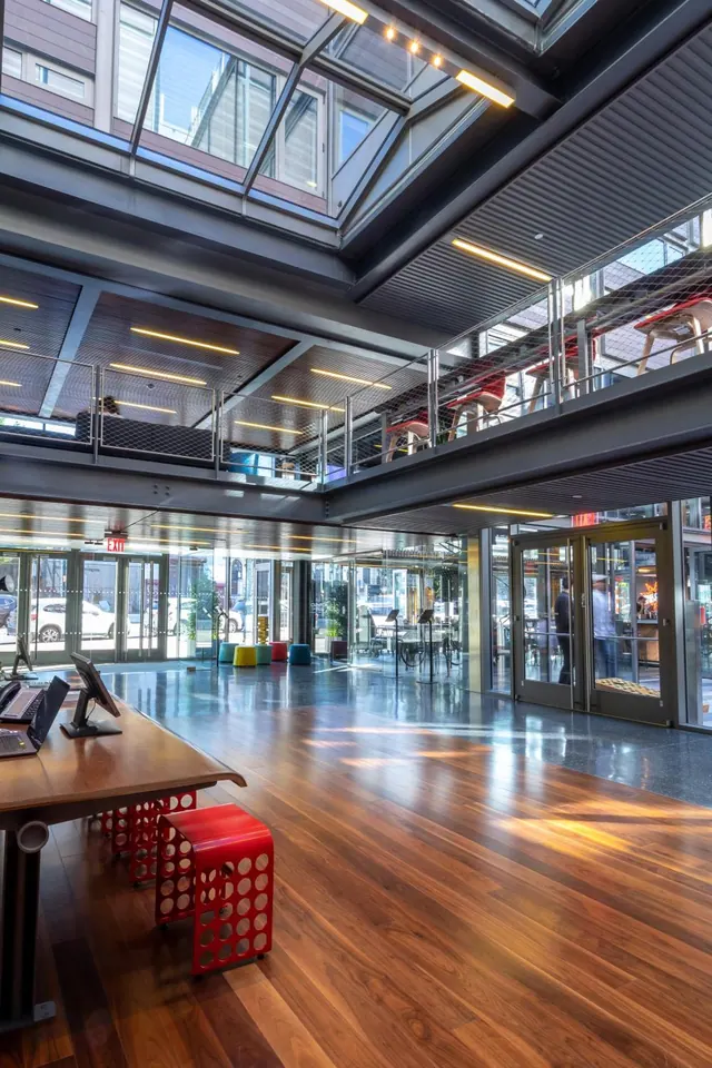 The photo shows a lobby with a wooden floor, a desk with computer monitors and red stools, a glass atrium overhead, and glass doors opening onto the street.