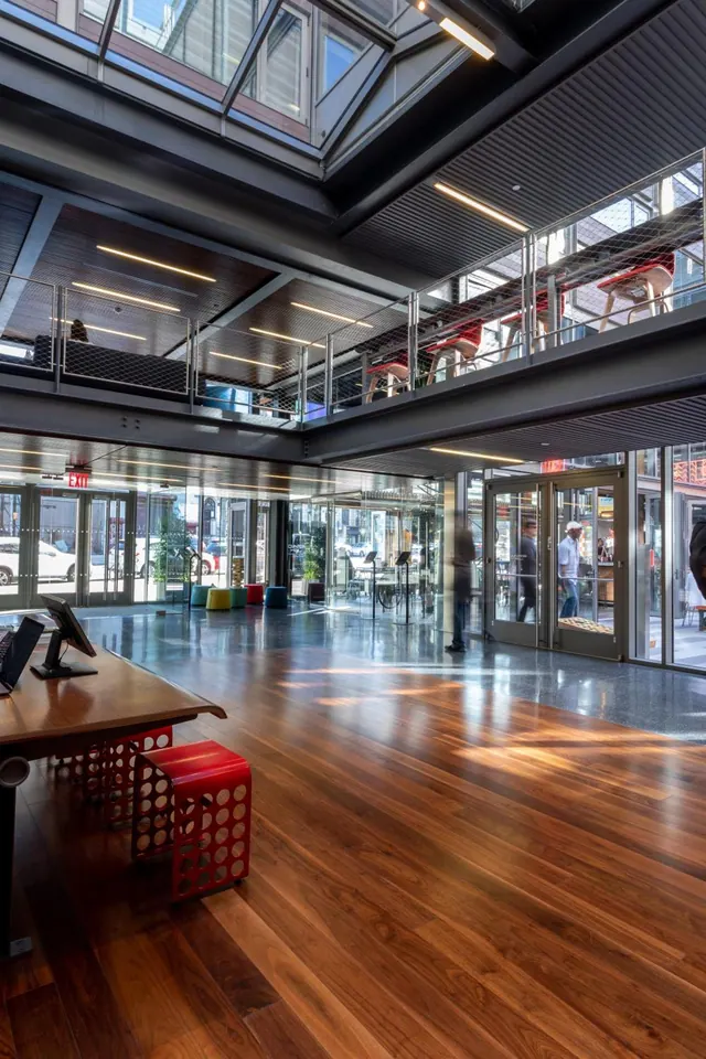 A lobby with a wood floor, a desk with a monitor, red stools on a mezzanine level above, and a few people walking near glass doors.