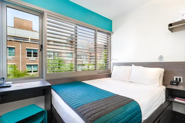 A bedroom with a double bed under teal and grey bedding, a desk by a window looking out onto a brick building and trees.