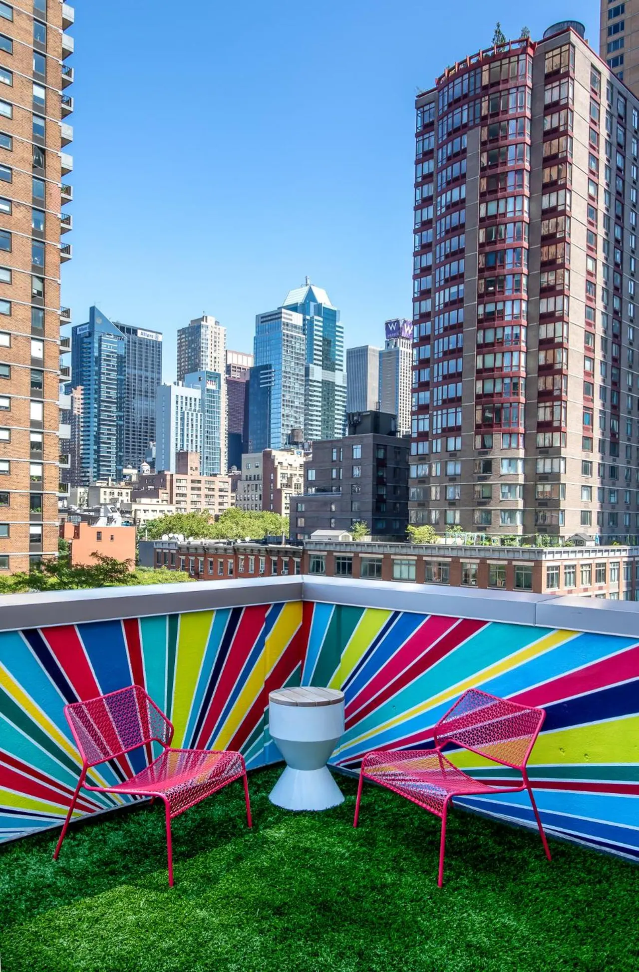 A rooftop terrace with a colorful striped wall, two red mesh chairs, a white side table and artificial grass, overlooking tall city buildings under a clear blue sky.
