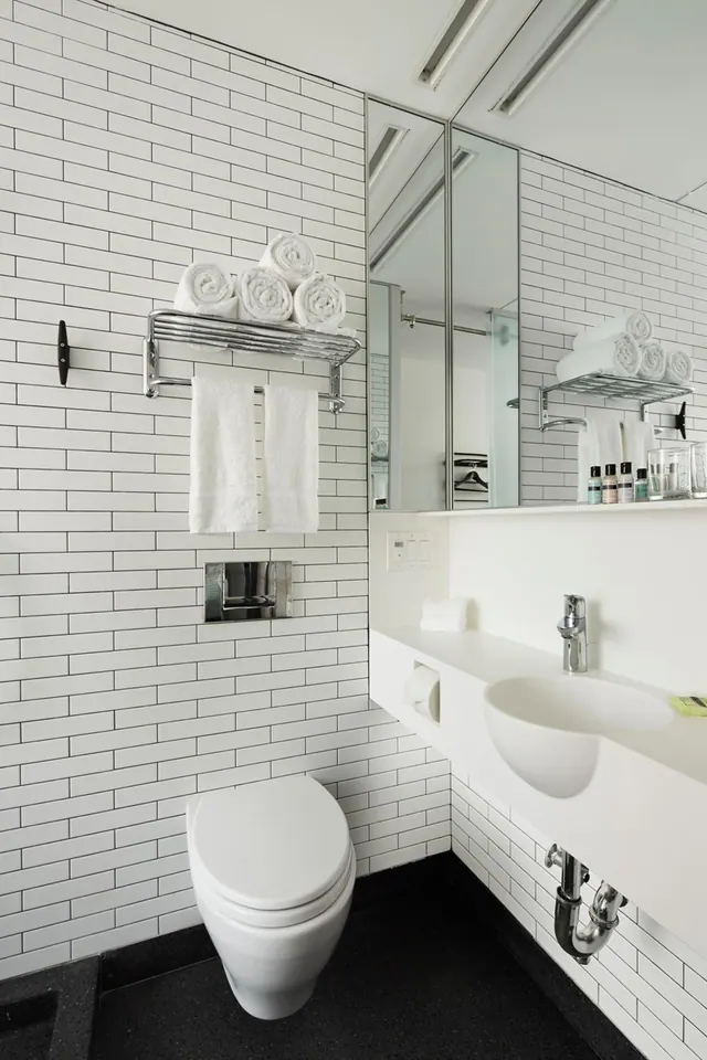 A bathroom with white subway tiles, a wall-mounted toilet, a round basin sink, rolled towels on a chrome shelf, and a large mirror.