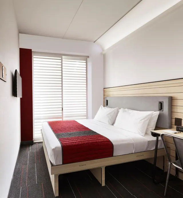 A small hotel bedroom with a made double bed with a red runner, a wall-mounted television, a window with white blinds, and a desk with a chair.