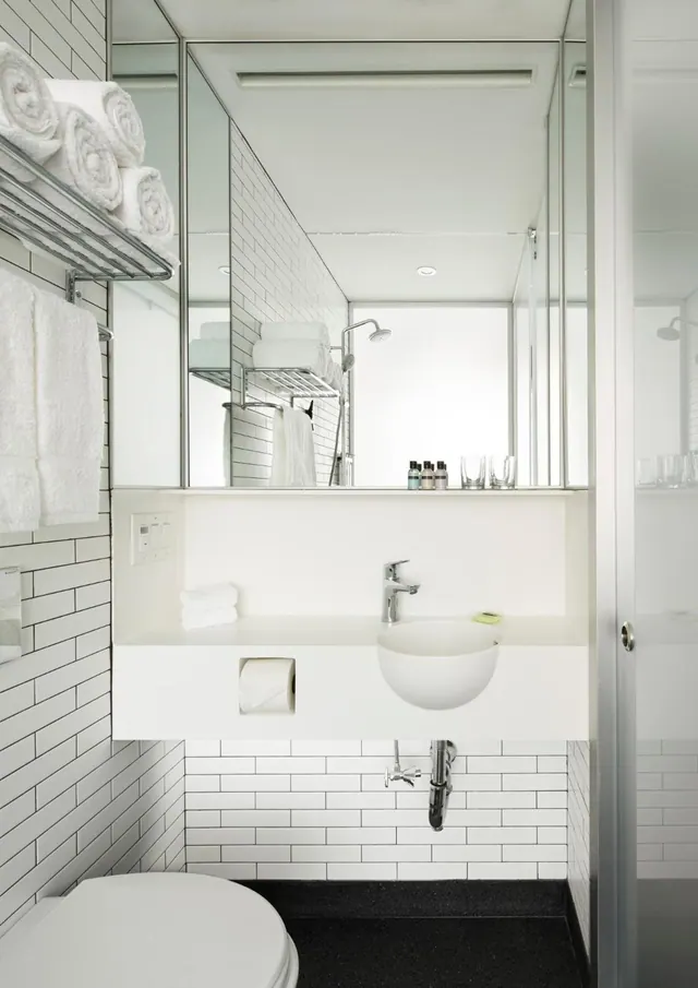 A white-tiled bathroom with a wall-mounted basin, a mirrored wall, towels on a wall shelf, a toilet in the foreground and a glass-enclosed shower on the right.
