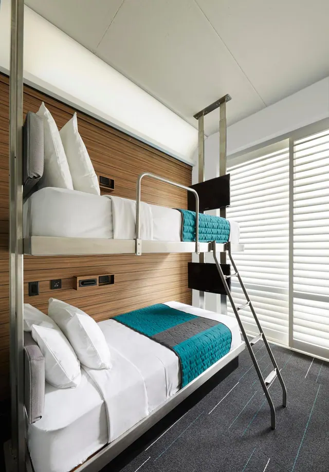 A bedroom with a stacked bunk bed against a wooden panelled wall, a metal ladder and rail, and white window blinds beside it.
