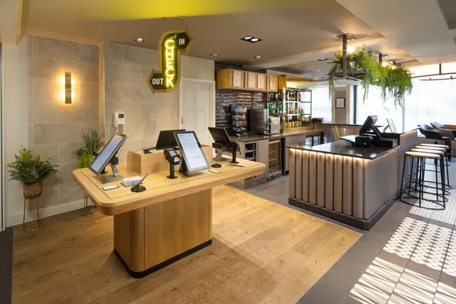 A hotel reception area with a wooden check-in desk, tablet terminals, a lit check-in and check-out sign, and a bar with stools in the background.