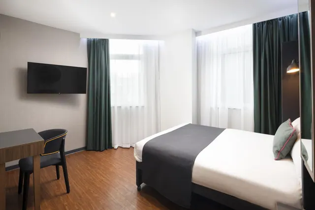 A double bed with a dark throw and grey cushion sits beside a wall-mounted television and a wooden desk with a chair, in a room with tall windows and green curtains.