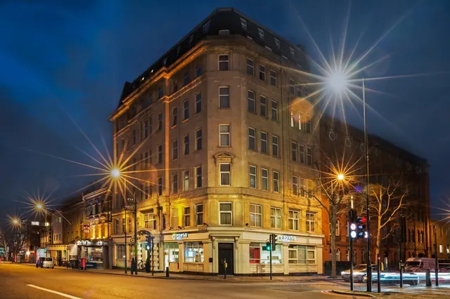 Night view of a corner stone building with illuminated ground-floor frontages, a Point A Hotel sign, and streetlights on the road in front.
