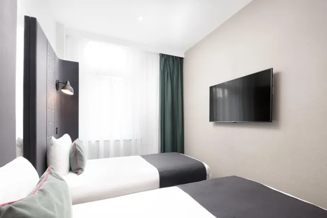A twin-bedded hotel room with a wall-mounted flat-screen television and sheer white curtains at the window.