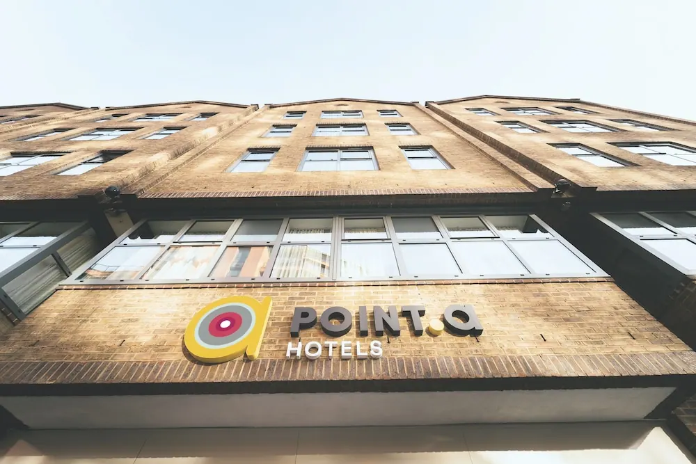 Low-angle view of a brick hotel building with a Point A Hotels sign above the entrance and rows of windows under a pale sky.