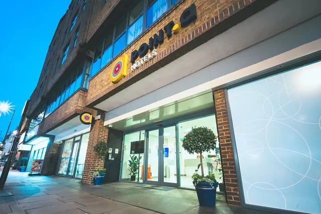 Exterior of a brick hotel building at dusk with illuminated Point A Hotels signage above glass entrance doors and potted trees on the pavement.