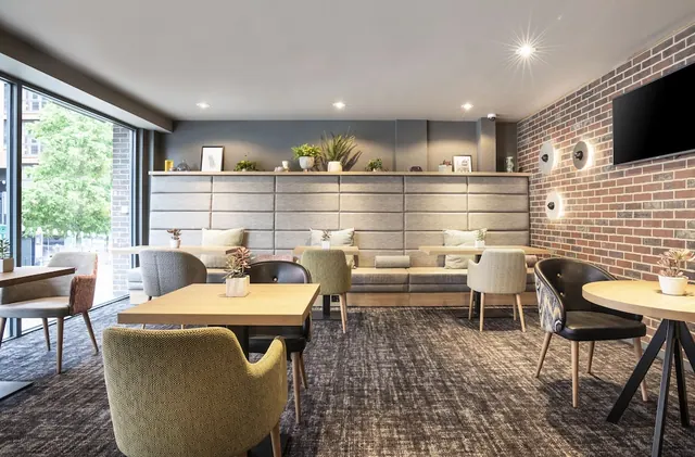 A lounge area with upholstered armchairs and small wooden tables, a padded banquette along the back wall, a brick feature wall with a wall-mounted television, and a large window on the left.