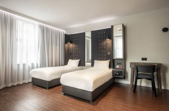A twin bedroom with two single beds with white linens and dark upholstered headboards, a desk with a chair on the right, and a window with sheer curtains on the left.