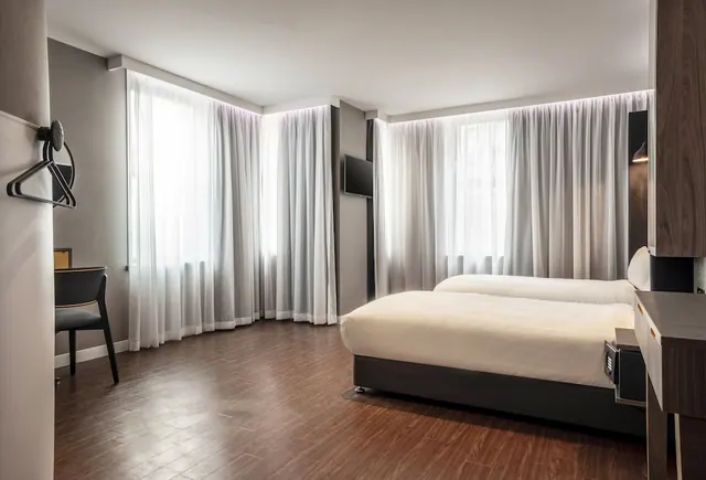 A hotel bedroom with two single beds, a wall-mounted television, a wooden desk and a chair, with sheer curtains covering the windows.