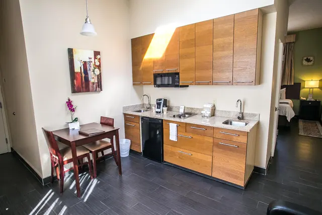 A small kitchenette with wooden cabinets, a sink, a microwave, a black mini fridge and a coffee maker, beside a small dining table with two chairs, with a bed visible through the doorway.