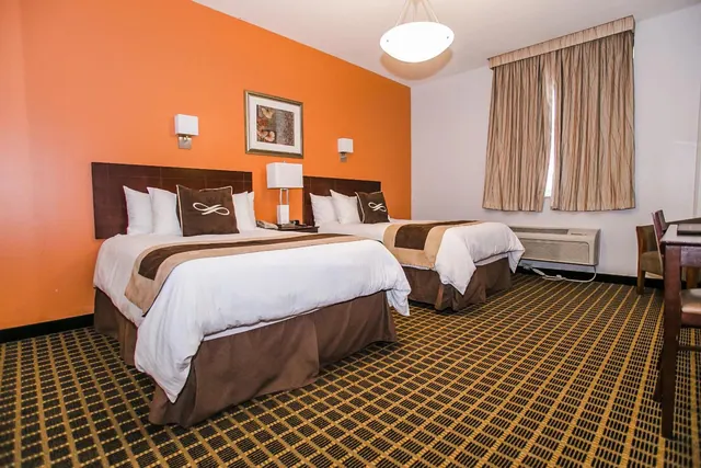 A hotel bedroom with two beds dressed in white linens, an orange accent wall, a wall air conditioner unit under the window and a desk on the right.