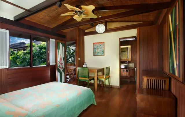 A bedroom with wood-panelled walls and ceiling, a ceiling fan, a bed with a green patterned cover, a small dining table with two green chairs, and windows looking out onto palm trees and a roof.