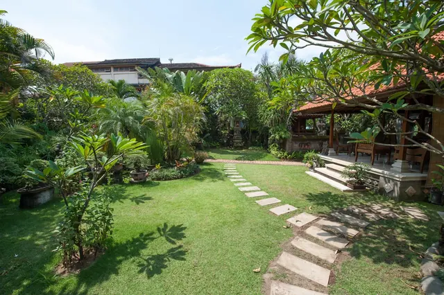 A grassy garden with a stepping-stone path leads toward a covered Balinese-style pavilion with a terracotta roof, surrounded by tropical plants and trees.