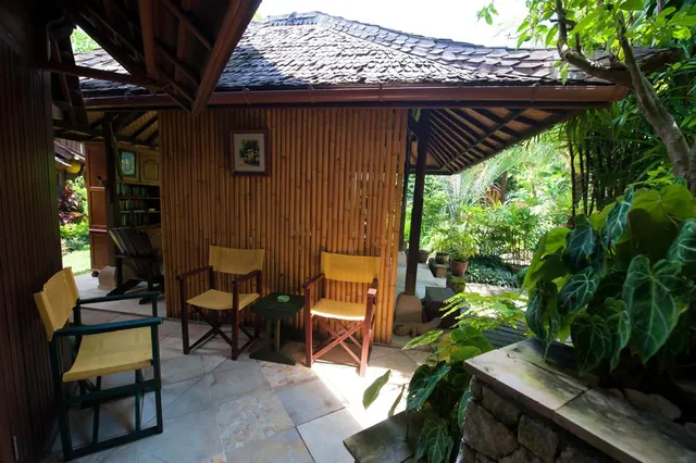 A covered terrace with a bamboo wall, two yellow director's chairs, a small side table and a stone tiled floor, with a tropical garden beyond.