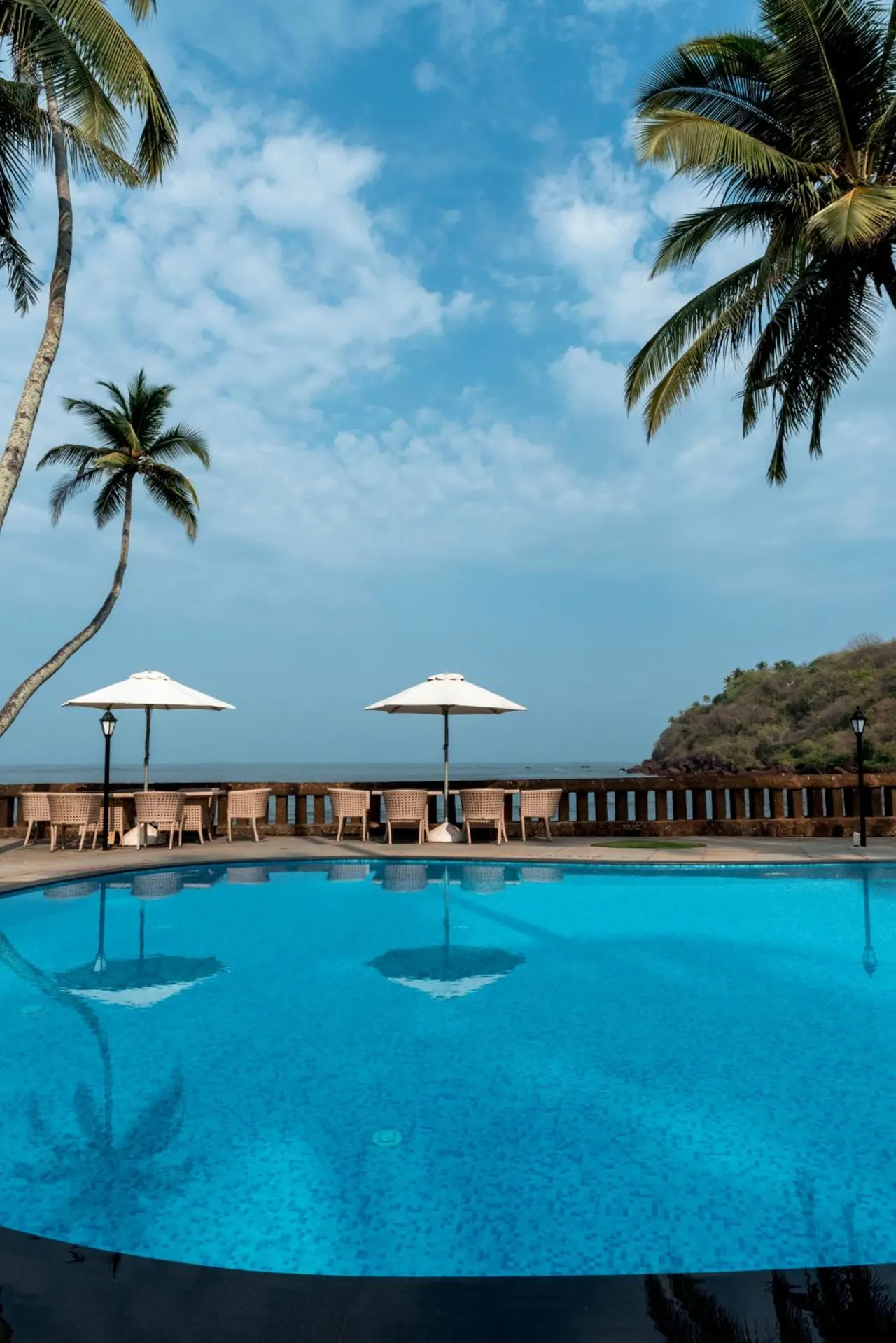 A swimming pool overlooks the sea with palm trees, umbrellas, and outdoor seating.