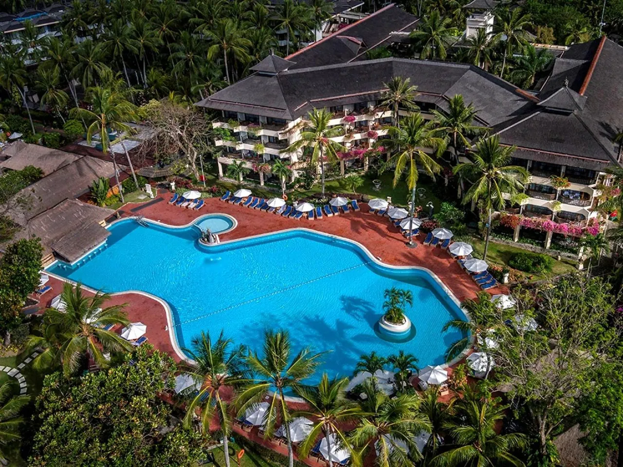 Aerial view of a large freeform swimming pool with sun loungers, white umbrellas, a small round pool and palm trees beside a multi-storey resort building.