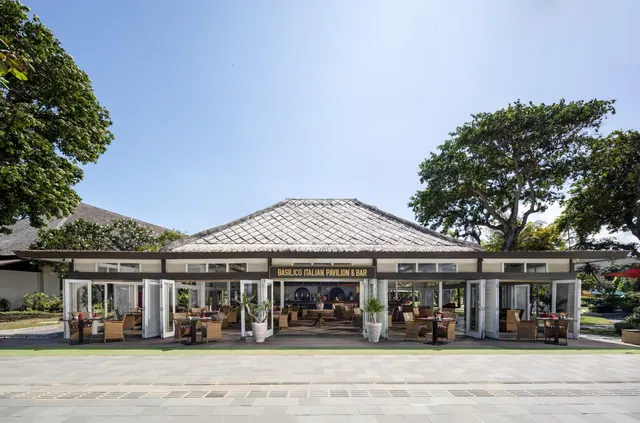 The front of the Basilico Italian Pavilion & Bar, a thatched-roof open-sided building with tables set out under the pavilion and trees on either side.