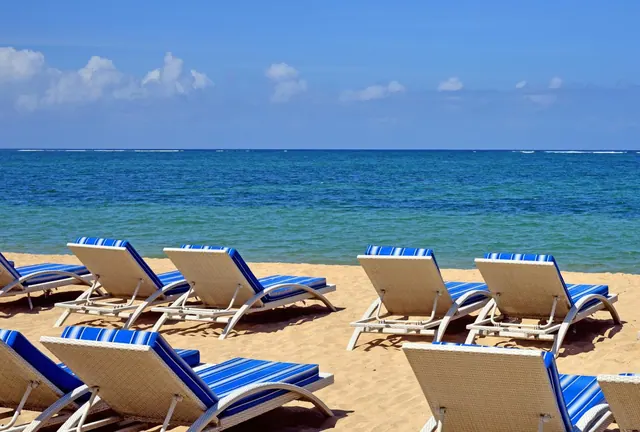 Rows of white wicker sun loungers with blue striped cushions sit on sand facing a calm blue sea under a partly cloudy sky.