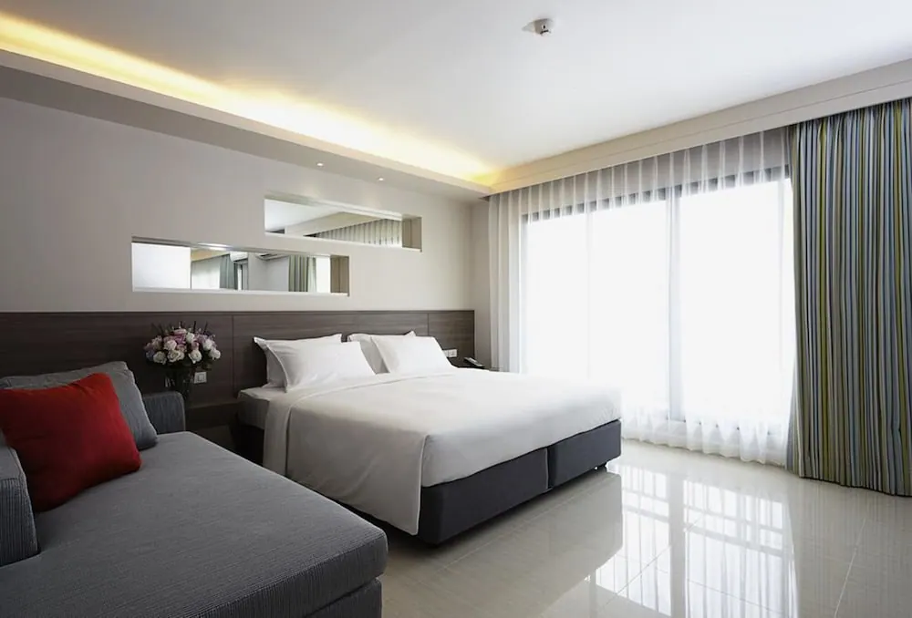 A bedroom with a made king bed, a grey sofa with a red cushion, sheer and striped curtains, and a large window letting in bright light.