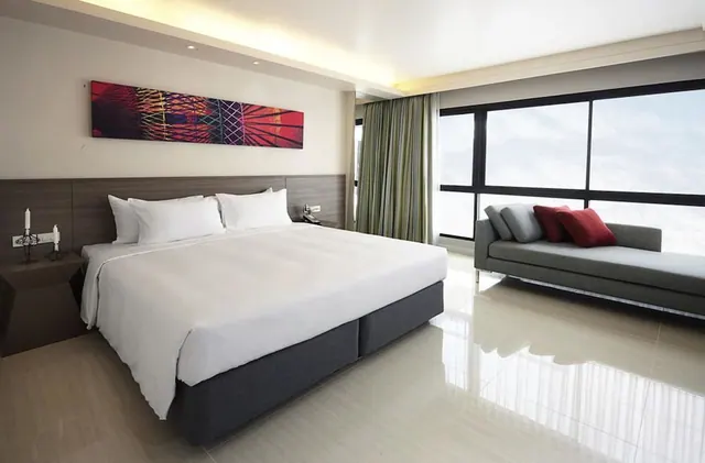 A bedroom with a king bed, a grey sofa with red cushions, and floor-to-ceiling windows along the right wall.