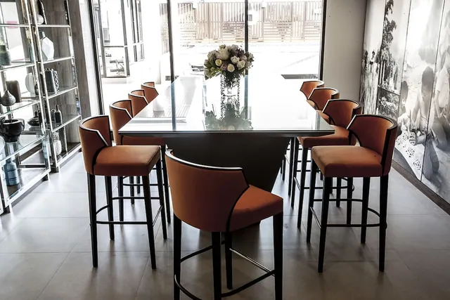 A high table with orange bar stools and a flower arrangement sits in a lounge beside tall glass doors and a black and white wall mural.