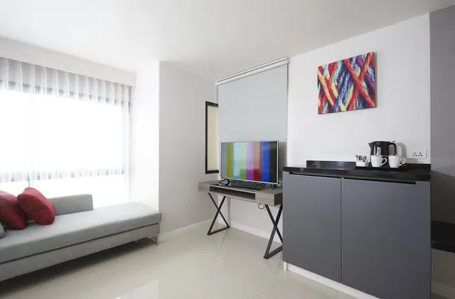 A modern living area with a grey daybed and red cushions, a television on a small desk, a wall painting, and a kettle and cups on a black-topped cabinet beside a bright window.
