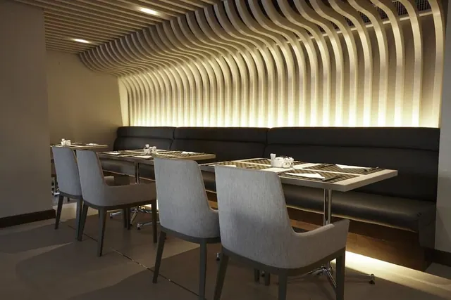 A restaurant dining room with set tables, grey upholstered chairs, a black leather banquette and a wavy wooden slatted wall lit from behind.