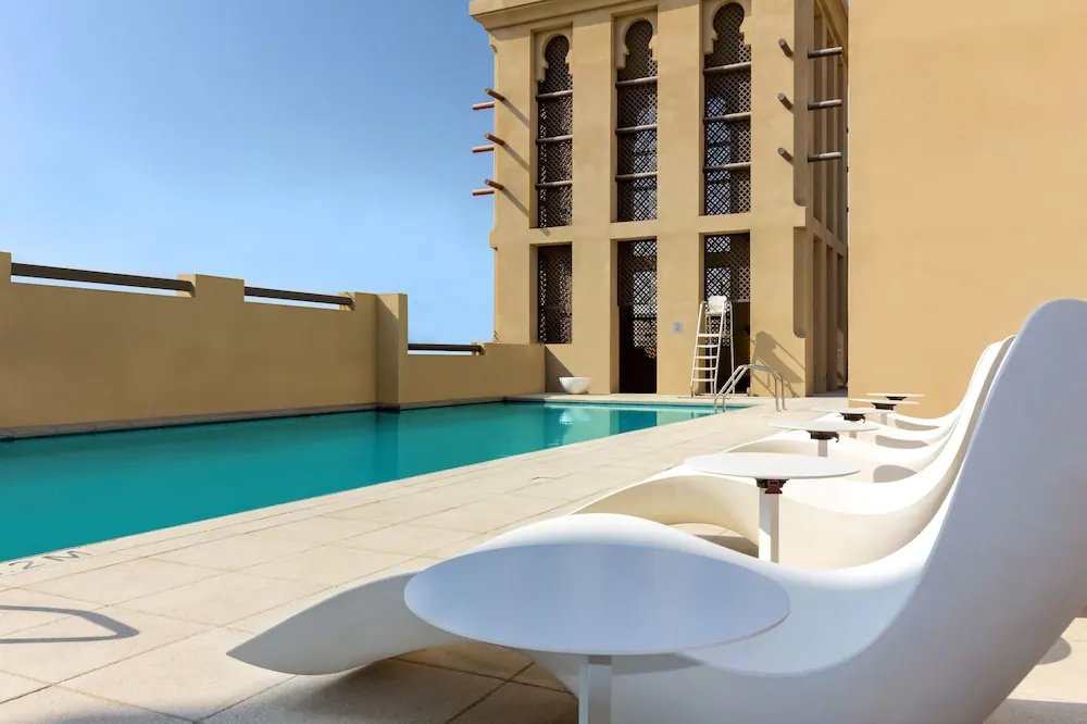 A rooftop swimming pool with white modern loungers and small side tables beside a beige building with arched latticed windows under a clear blue sky.
