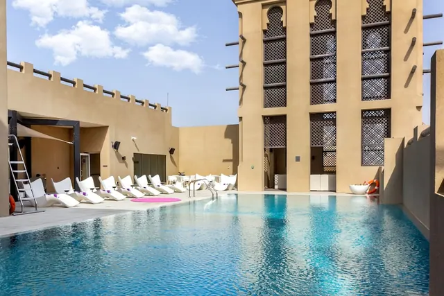 An outdoor swimming pool with white sun loungers beside a beige building with latticed traditional windows under a partly cloudy sky.