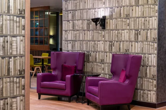 Two purple wingback armchairs and a small round side table stand in front of a bookshelf-patterned wall in a hotel seating area.