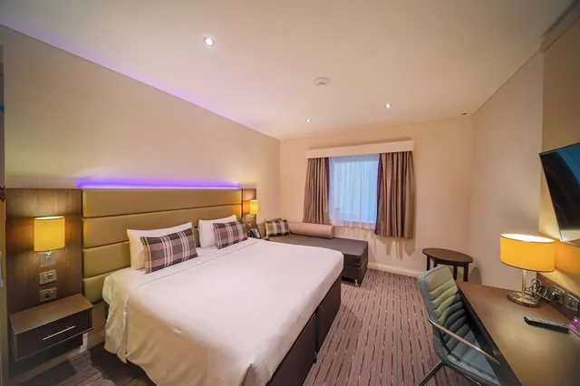 A hotel bedroom with a double bed, a daybed under the window, a wall-mounted television and a desk with a chair.