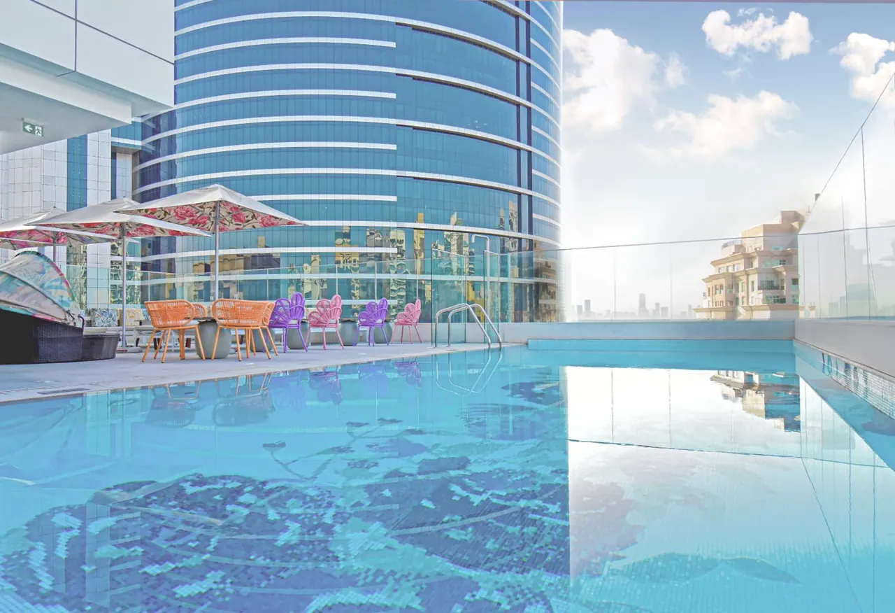A rooftop swimming pool with a glass railing, colourful chairs and parasols beside it, with a glass tower and city buildings in the background.