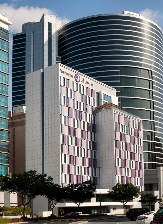 Exterior of a Premier Inn hotel building with lilac window accents, set between tall glass office towers with trees and parked cars along the street.