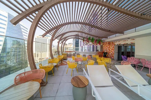 A rooftop terrace under a curved wooden pergola with yellow and pink woven chairs, white sun loungers, and a view of high-rise buildings beyond the glass railing.