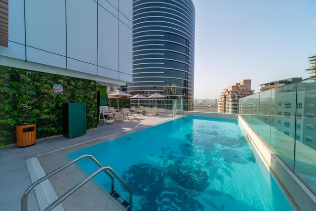 A rooftop swimming pool with glass railings, sun loungers and shade umbrellas, with a curved tower and residential buildings beyond.